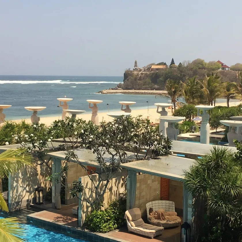 A beachfront resort in Bali, pool cabanas and stone urns above the sand, a clifftop temple beyond.